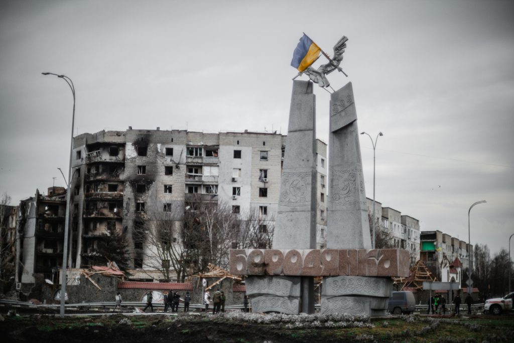 Damaged buildings and a structure with the Ukrainian flag in a war zone, with people and scattered debris in the foreground.