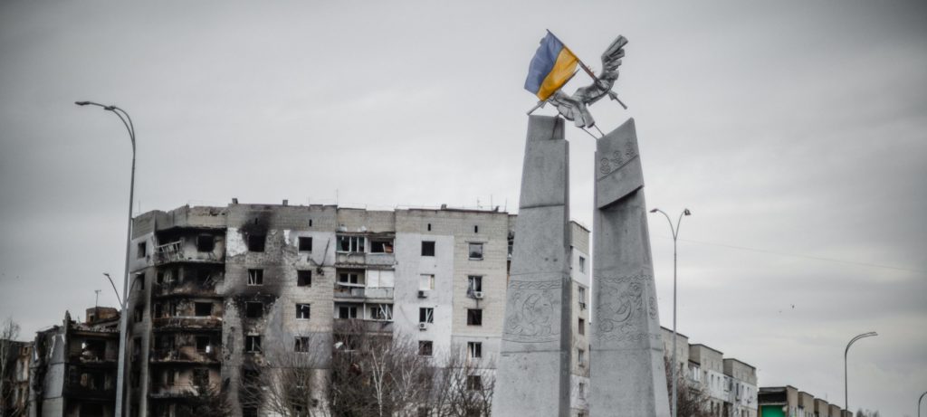 Damaged buildings and a structure with the Ukrainian flag in a war zone, with people and scattered debris in the foreground.