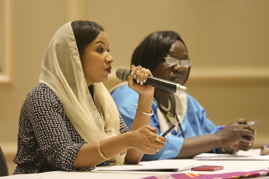 Two black women sit at a table during a formal meeting. One speaks into a microphone while the other listens attentively with a pen in hand.