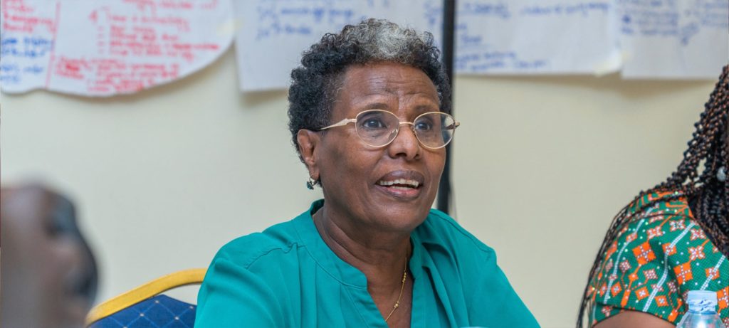 Elderly black woman in a green shirt speaks during a discussion meeting with notes and water bottles on the table.