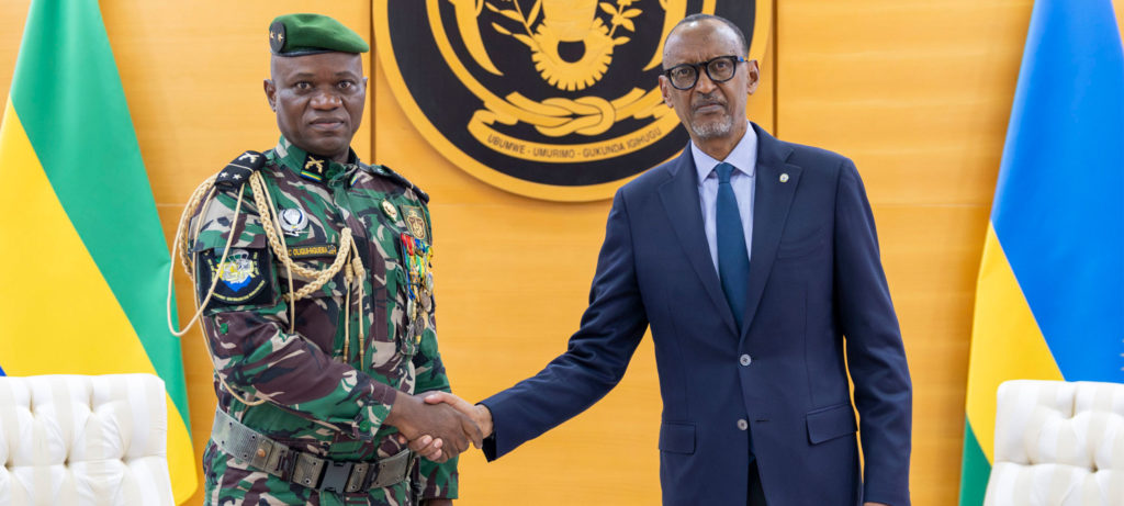 General Oligui Nguema, the current President of Gabon on the left shaking hands with another man in front of an official seal, dressed in a military uniform and a suit, with flags in the background.