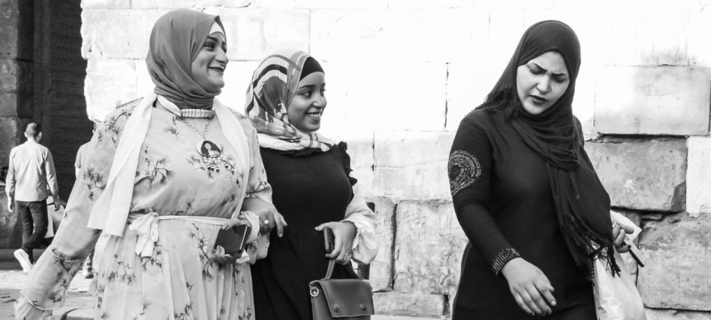 Black and white photo of three women in hijabs walking together on a cobblestone street, two smiling and chatting while the third looks down thoughtfully.