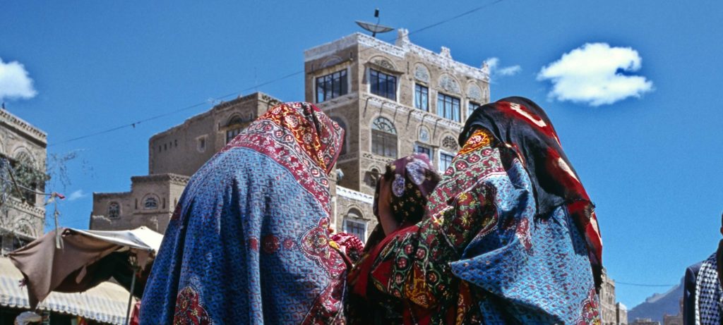 Two women wearing colorful patterned shawls stand closely together at an outdoor market, with traditional buildings and blue sky in the background.